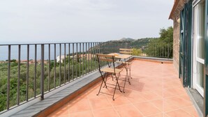 Een balkon met een tafel en stoelen, een uitzicht op zee en een bergachtig landschap op de achtergrond.