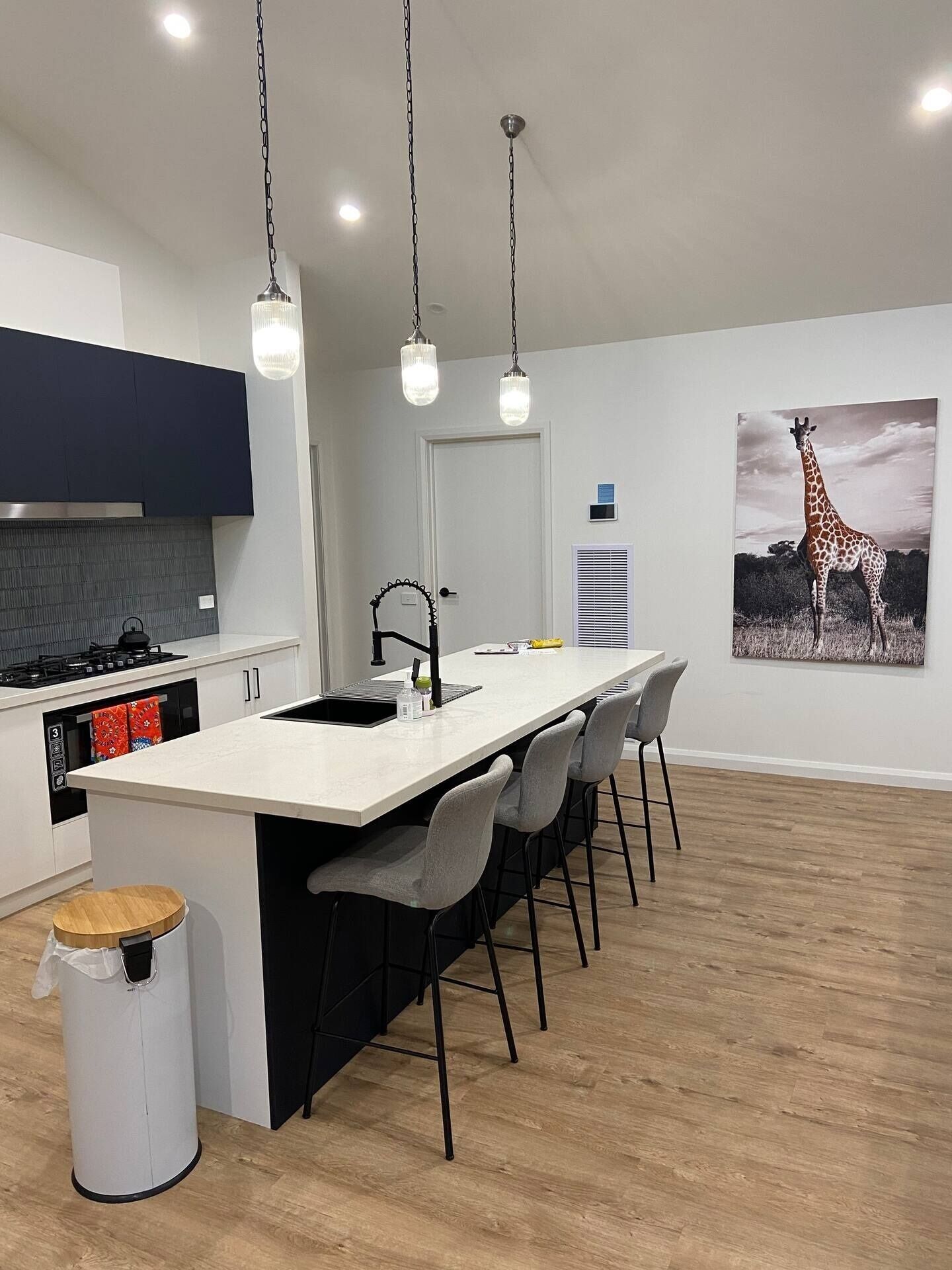 Una cocina moderna con una isla central, taburetes de bar, un cubo de basura y un cuadro de una jirafa en la pared.