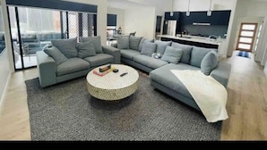 Een moderne woonkamer met een hoekbank, een ronde salontafel en een groot raam dat natuurlijk licht binnenlaat.