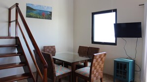 Ein Zimmer mit einer Holztreppe, einem Esstisch mit Stühlen, einem an der Wand montierten Fernseher und einem Fenster.