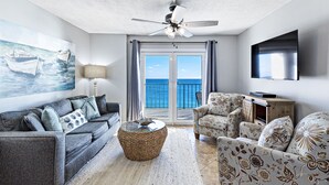 Ein Wohnzimmer mit einer Couch, einem Sessel und einem runden Couchtisch. Es gibt einen Deckenventilator, ein Gemälde mit Booten und Meerblick durch die Glastüren.