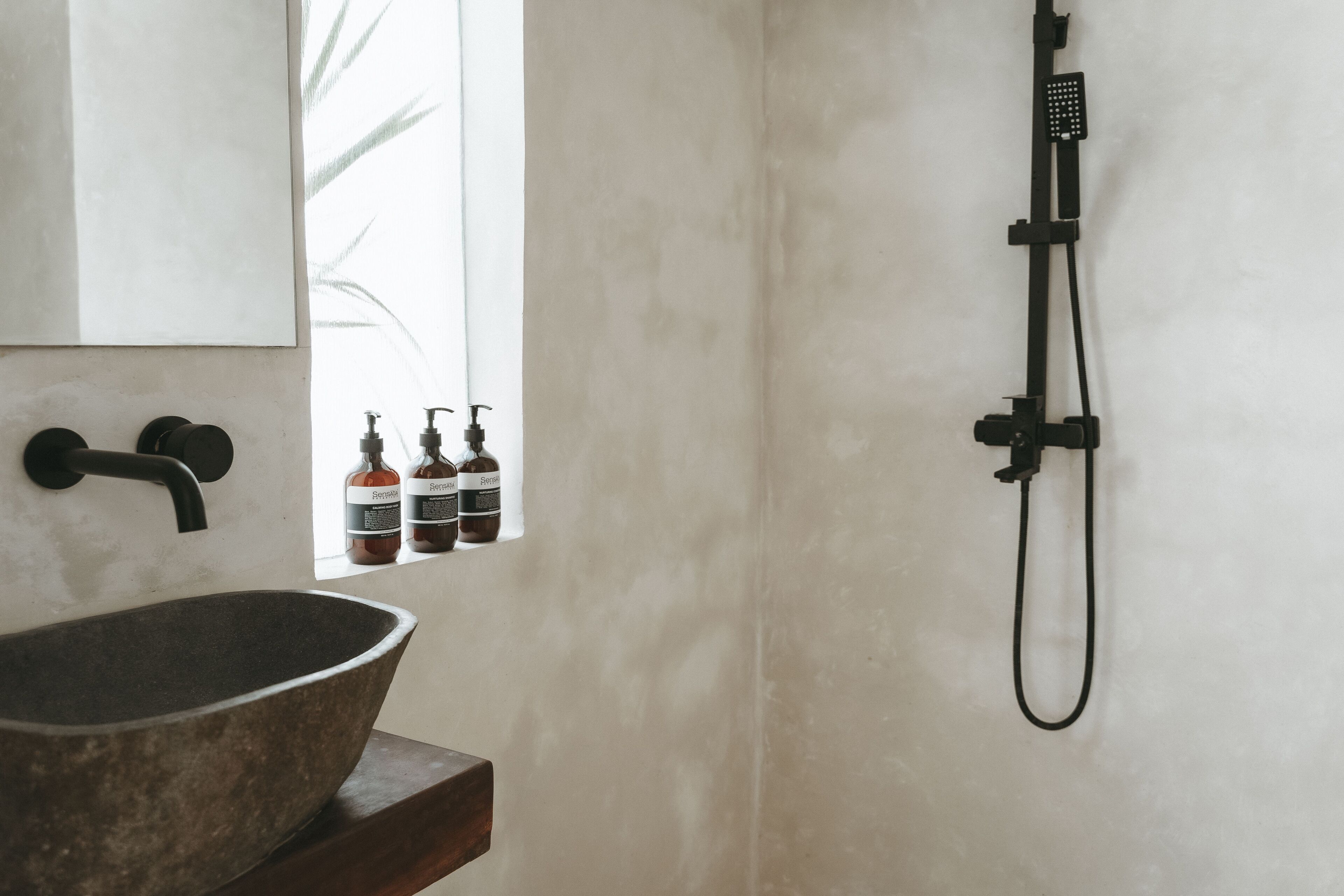A bathroom with a large stone basin, a black faucet, a handheld showerhead, and a window with bottles of shampoo and conditioner.