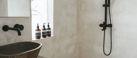 A bathroom with a large stone basin, a black faucet, a handheld showerhead, and a window with bottles of shampoo and conditioner.
