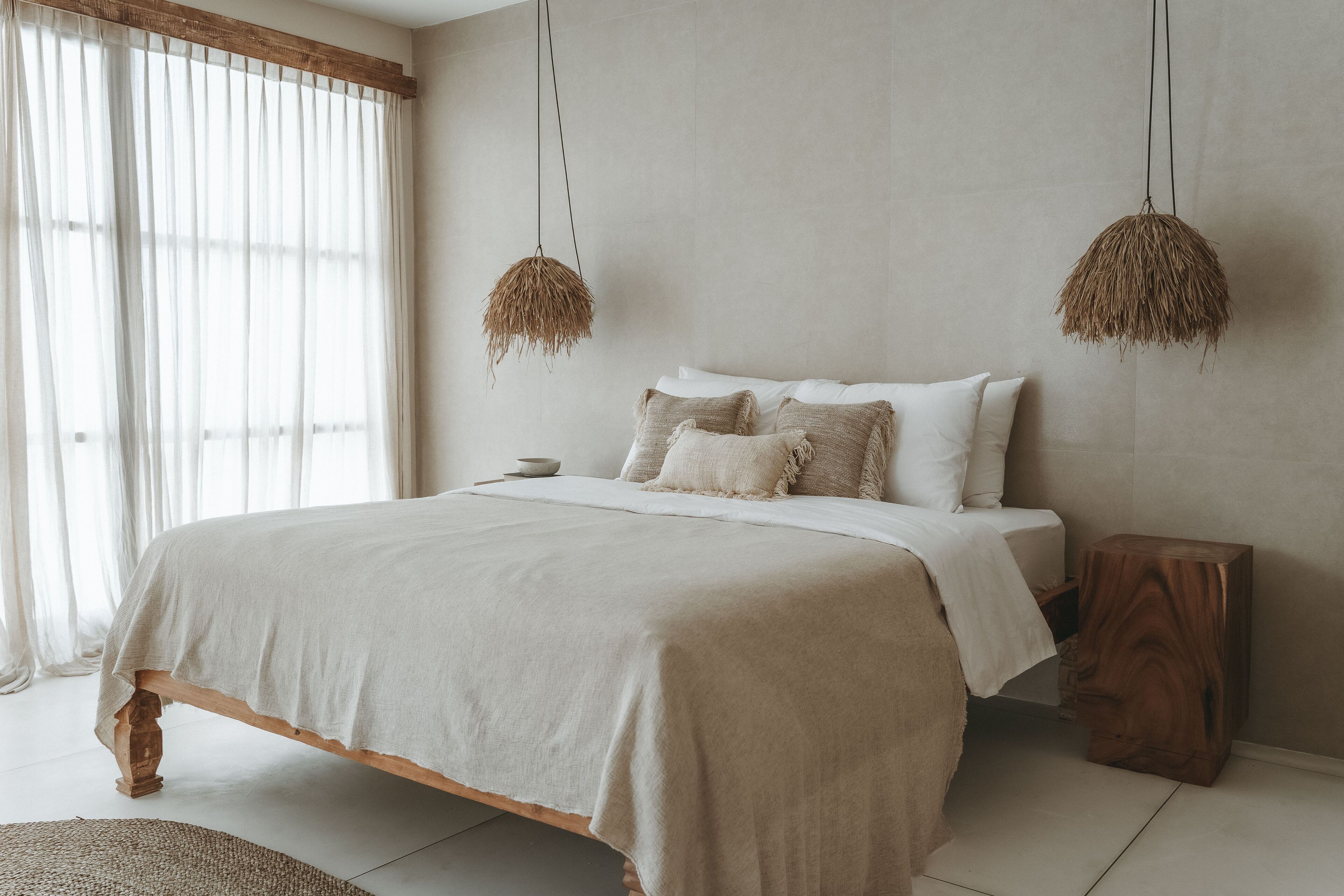 A bedroom with a large bed, two hanging light fixtures, a bedside table, and a window with sheer curtains.
