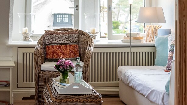 Ein gemütliches Zimmer mit einem Korbstuhl, einer gepolsterten Bank, einem Nachttisch mit Lampe und Blick ins Freie.