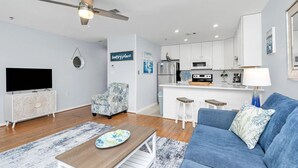 Una sala de estar moderna con un sofá, una mesa de centro, un televisor y una cocina con refrigerador, microondas y una pequeña isla.
