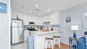 A modern kitchen with white cabinets, a stainless steel refrigerator, a microwave, and a stove. There are two bar stools and a trash bin.