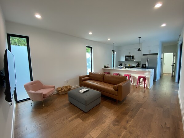 Un salon moderne avec un canapé en cuir marron, un pouf gris et un fauteuil rose. Un téléviseur à écran plat est fixé au mur.