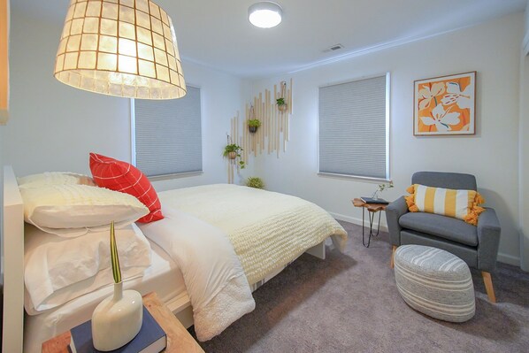 A bedroom with a bed, a chair, a side table, a lamp, and a window with blinds.