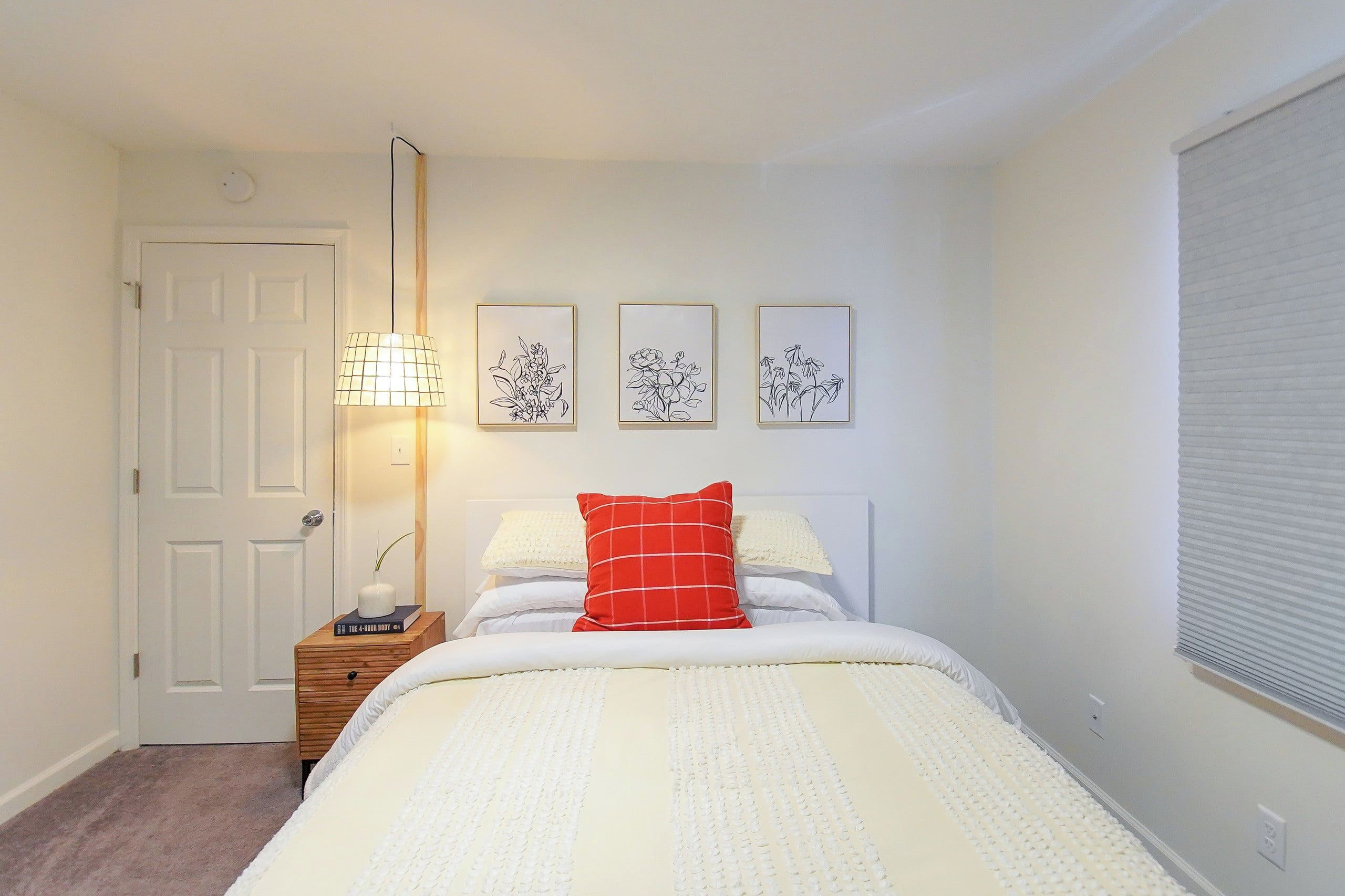 Un dormitorio con cama, mesita de noche, lámpara, puerta y tres cuadros enmarcados.