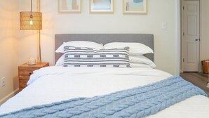 Un lit bien fait, recouvert d’une couverture bleue, une lampe de chevet et trois œuvres d’art encadrées accrochées au mur.