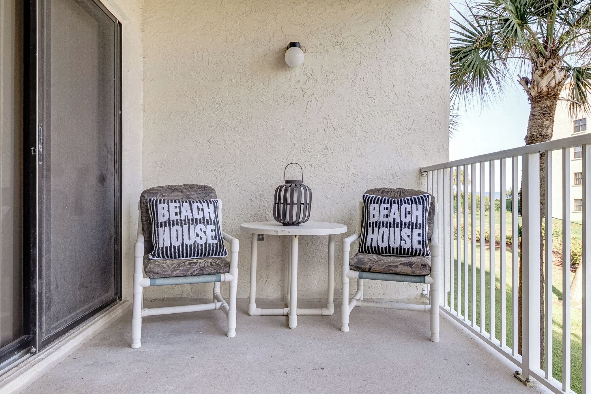 A balcony with two chairs and a small table, each with a pillow labeled "Beach House".