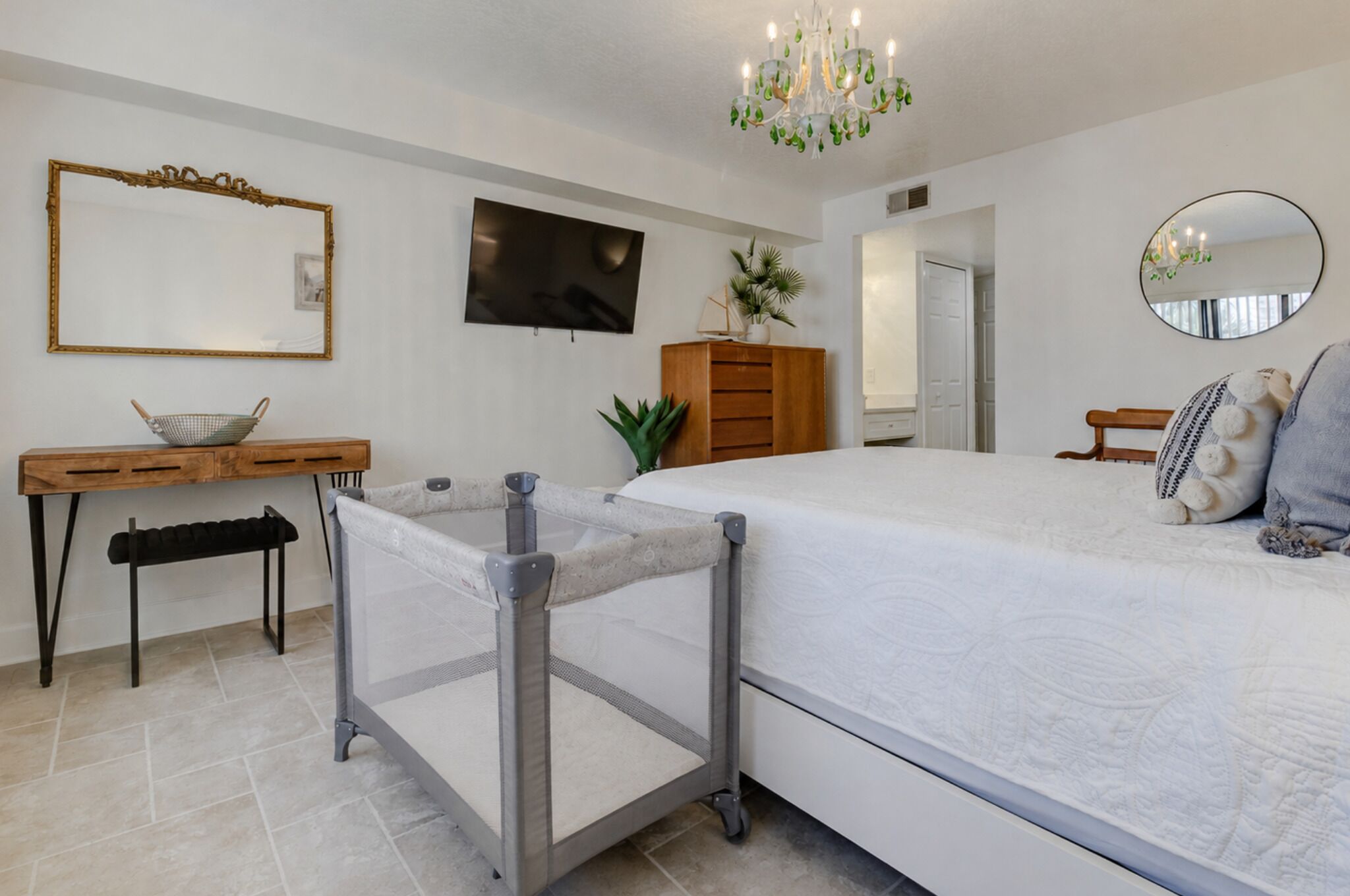 A bedroom with a bed, a desk, a chair, a television, a mirror, and a chandelier.