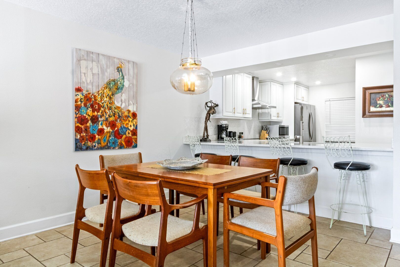 A dining area with a wooden table and chairs, a hanging light fixture, and a colorful painting on the wall.