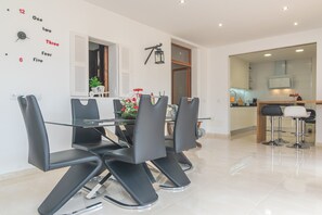 A modern dining area with a glass table and black chairs, a kitchen visible through an open door, and a wall clock.