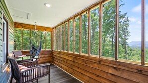Une terrasse en bois avec un fauteuil suspendu et une table, de grandes fenêtres offrant une vue sur les arbres et les montagnes.