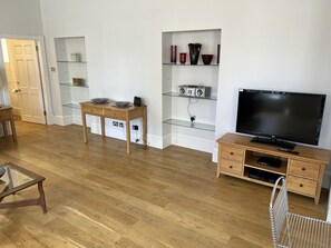 A modern living room with a wooden floor, a glass-top coffee table, a TV stand with a flat-screen TV, a bookshelf with decorative items, and a door leading to another room. - Historic Luxury Apartment in Inverness City Centre (Inverness)
