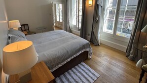 Un dormitorio con una cama grande, una mesita de noche, una lámpara y vistas a la ciudad desde las ventanas.
