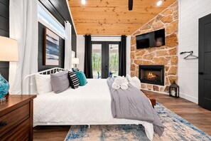A bedroom with a bed, a nightstand, a TV mounted on a stone wall, and a fireplace.
