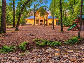 A cabin with a large porch, surrounded by trees and a playground.