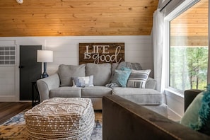 A cozy living room with a grey sectional sofa, a wooden sign saying "Life is good," and a large window offering a view of trees.