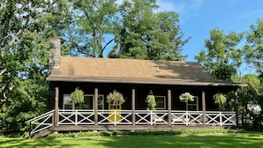 A rustic wooden cabin with a porch, surrounded by lush greenery and trees.