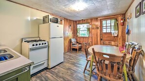 Eine kleine Küche mit Herd, Kühlschrank und Spüle. Ein Holztisch mit Stühlen und ein Fenster mit Vorhängen.