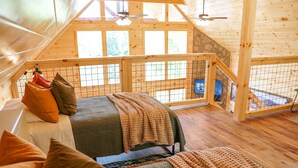 A cozy loft bedroom with a high ceiling, wooden beams, a bed with multiple pillows, a checkered throw blanket, and a view of the outdoors through large windows. - Perfect Family Cabin for 4–5 • Kids' Loft, Arcade, Hot Tub, Easy Drive (Gatlinburg)