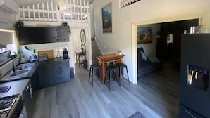 Una cocina compacta con estufa integrada, una zona de comedor con taburetes, una barandilla blanca en el nivel superior y un refrigerador.