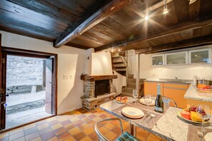 A rustic kitchen with a stone fireplace, a dining area with a table set for two, and a view of a stone wall through a glass door.