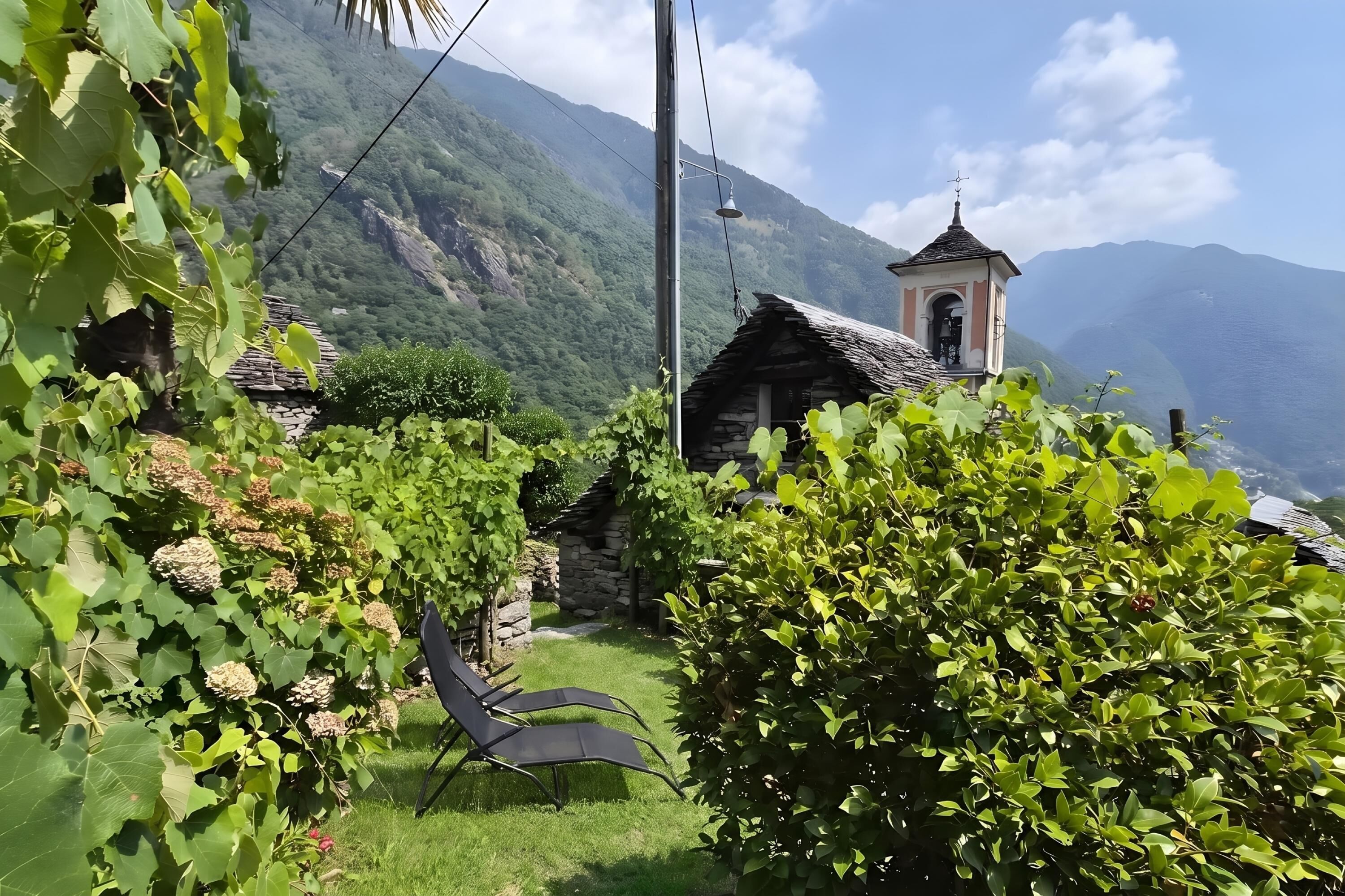 A stone building with a bell tower, surrounded by lush greenery and a mountainous backdrop.