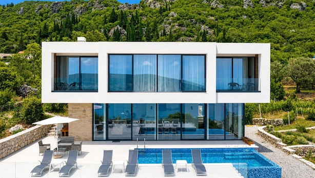 A modern two-story building with a swimming pool, outdoor seating, and a view of a mountainous landscape.