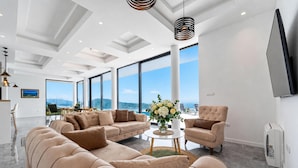A spacious living area with large windows, a sofa set, a coffee table, and a flat-screen TV mounted on the wall.