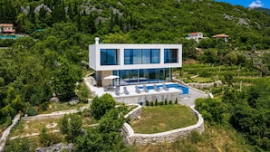 A modern two-story house with a swimming pool, surrounded by greenery and mountains.