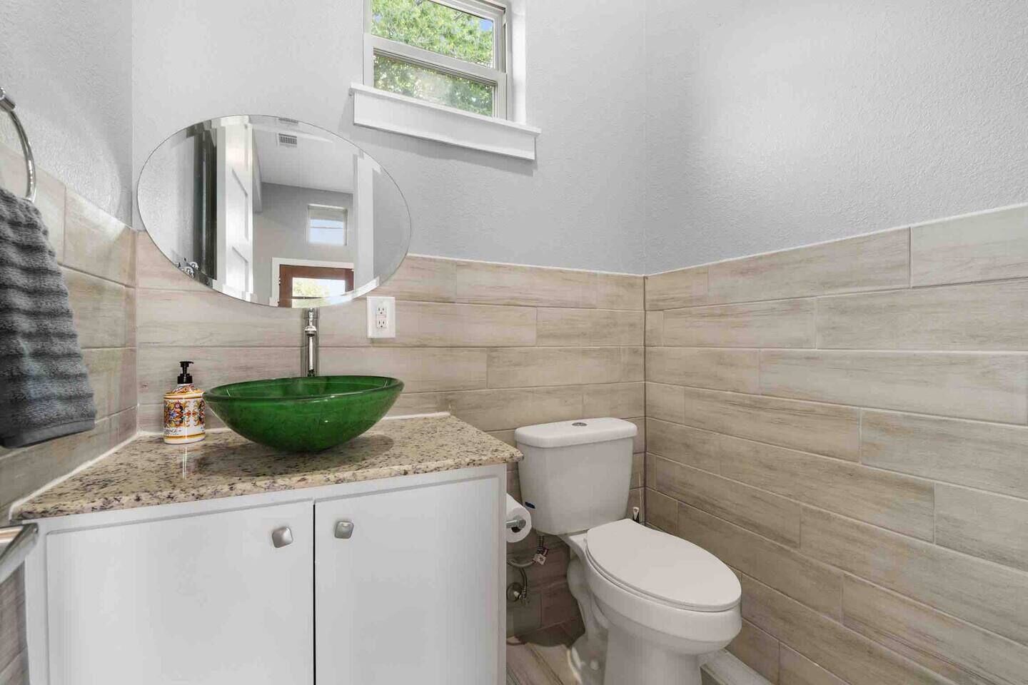 A bathroom with a green bowl sink, white toilet, and a window above the sink.