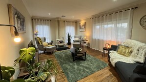 Ein Wohnzimmer mit einer Couch, einem Sessel, einem Couchtisch und einem Kamin. Es gibt Blumentöpfe und eine Uhr an der Wand.