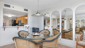 A dining area with a glass table surrounded by wicker chairs, a kitchen in the background, and a large mirror reflecting the space. - Oceanfront Condominium with Swimming Pool. (Saint Simons Island)