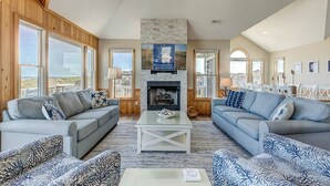 A spacious living room with two sofas, a coffee table, and a fireplace. Large windows provide views of the outdoors. - What the Shell | Oceanfront | Private Pool, Hot Tub, Community Pool (Corolla)