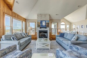 A spacious living room with two sofas, a coffee table, and a fireplace. Large windows provide views of the outdoors. - What the Shell | Oceanfront | Private Pool, Hot Tub, Community Pool (Corolla)