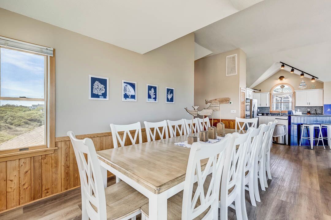 A dining area with a wooden table and white chairs, a kitchen with white cabinets, and a view of greenery outside.