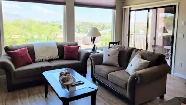 A living room with two sofas, a coffee table, and a lamp. There are large windows and a sliding glass door leading to an outdoor area. - Beautiful Mountain Views and a spacious relaxing home in NW Tucson. (Tucson)