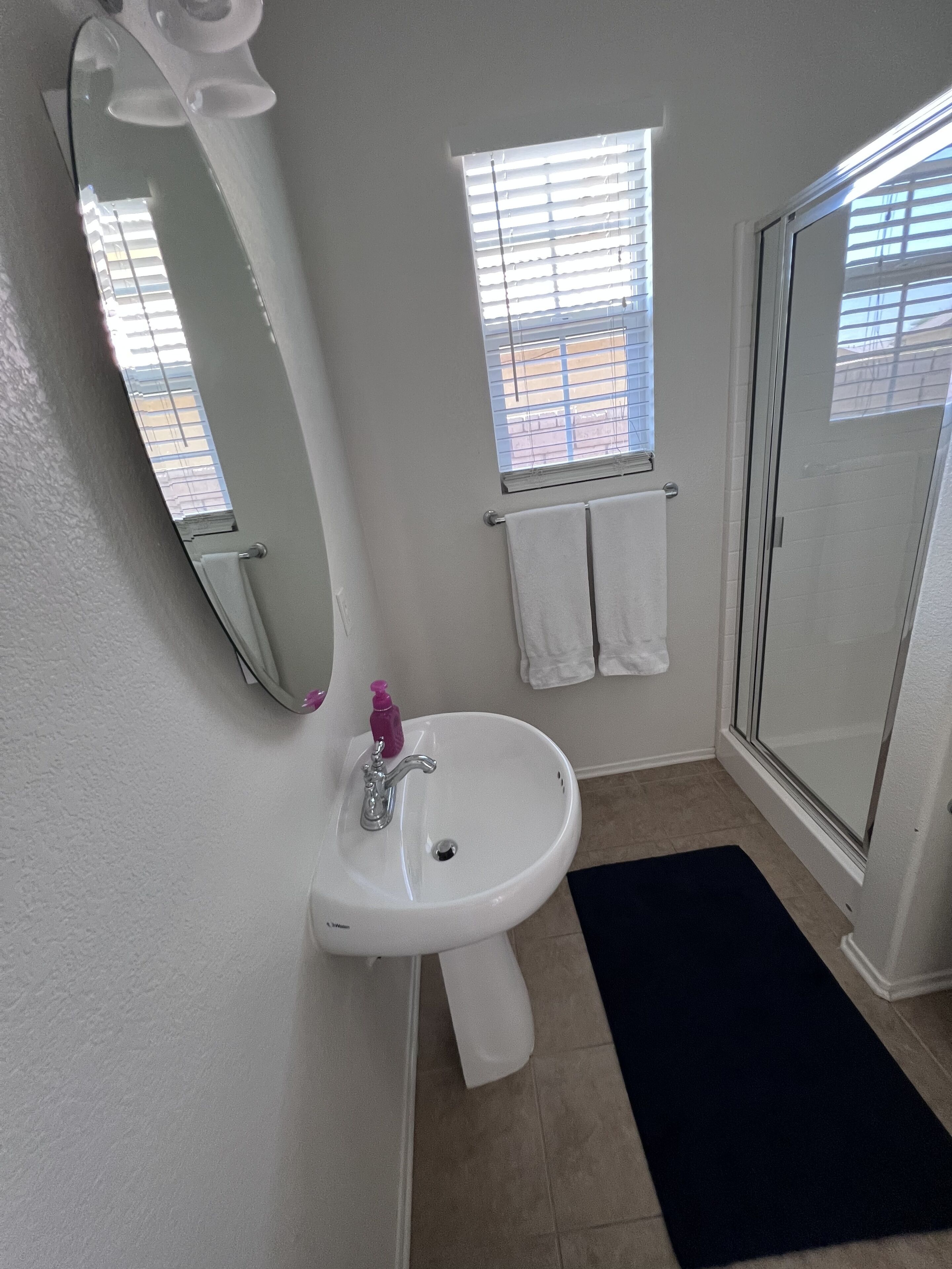 Un baño con un lavabo blanco, un espejo, una ventana con persianas y una puerta de cristal para la ducha.