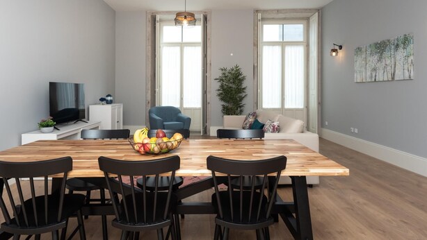 Ett modernt vardagsrum med ett matbord i trä, svarta stolar, en blå fåtölj, en vit soffa, en tv på ett vitt tv-bord och ett stort fönster med gardiner.