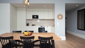 Una cucina moderna con mobili bianchi, un tavolo da pranzo in legno e sedie nere. Sul tavolo c'è una ciotola di frutta e sopra pendono delle luci a sospensione.