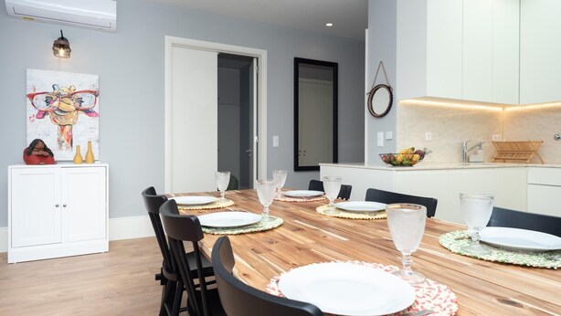 Un comedor moderno con una mesa de madera para seis personas, armarios blancos y una zona de cocina con frutero y fregadero.