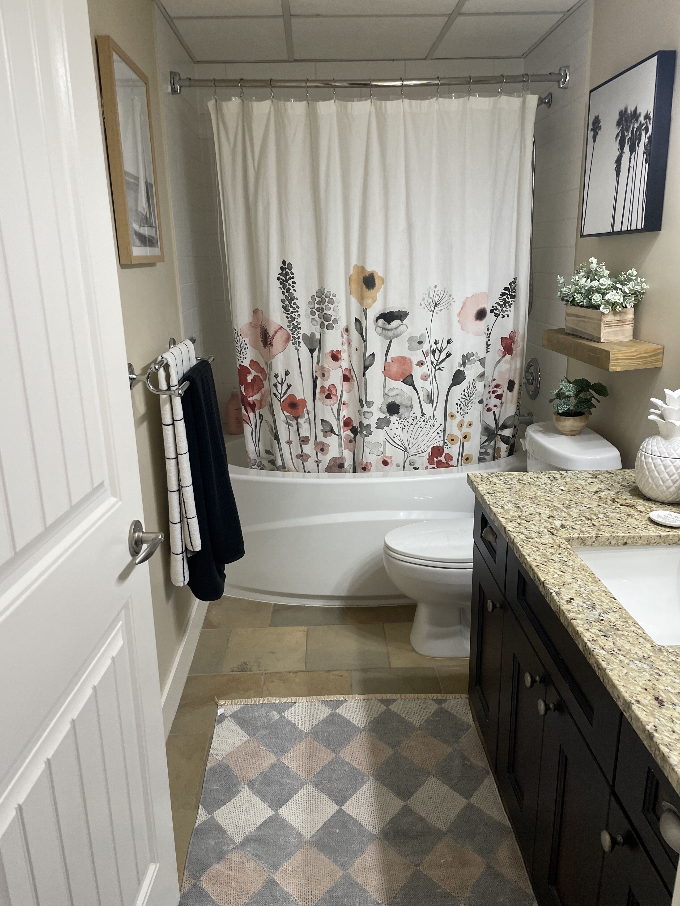 Une salle de bain avec un rideau de douche orné de motifs floraux, des toilettes, un lavabo avec un plan de travail en granit et un tapis à motifs.