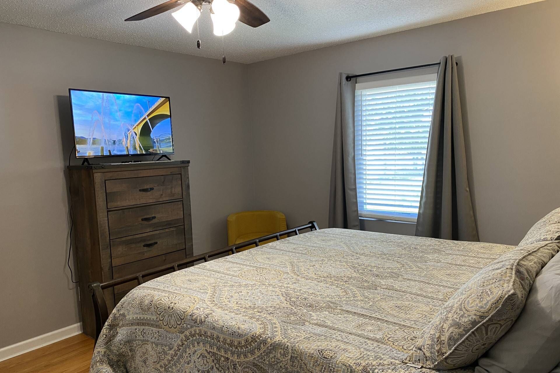 Ein Schlafzimmer mit einem Bett, einem Fernseher auf einer Holzkommode, einem Fenster mit Jalousien und einem Deckenventilator.