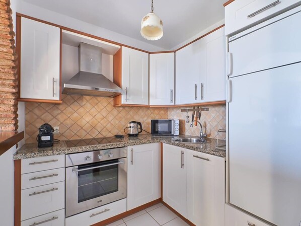 Cocina compacta con armarios blancos, horno de acero inoxidable, microondas y cafetera.