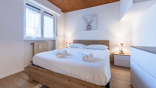 A bedroom with a wooden ceiling, a large bed, two bedside tables with lamps, a radiator, and a window with curtains. - One bedroom Apartment in Canazei (Campitello di Fassa)