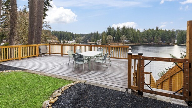 Eine Holzterrasse mit Tisch und Stühlen, mit Blick auf einen See, im Hintergrund Bäume und Häuser.
